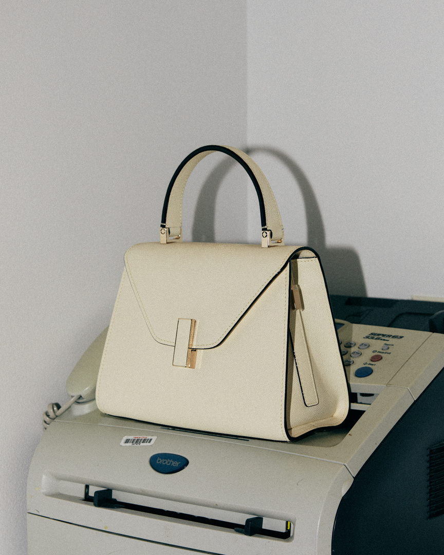 Cream colored leather Valextra handbag sits atop a light grey Brother printer against a pale grey studio wall backdrop.