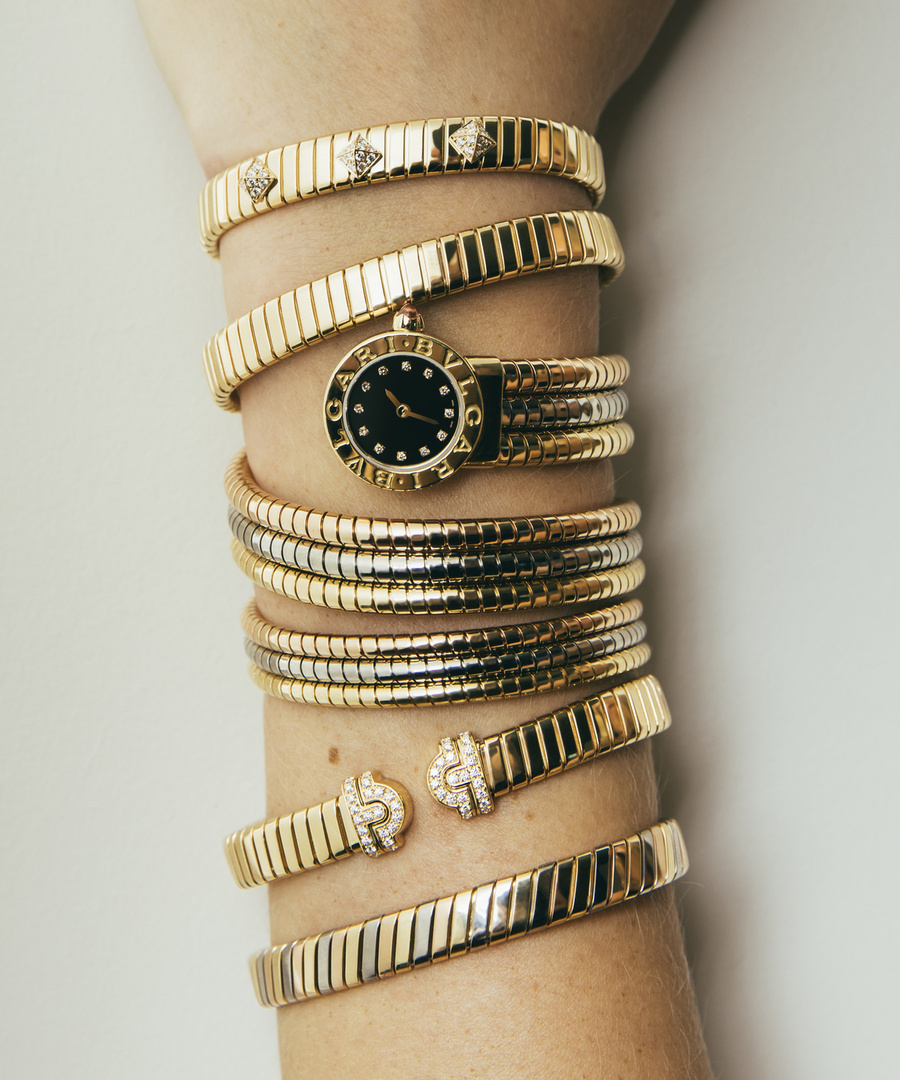 Close-up of a person's arm adorned with multiple gold Bulgari Serpenti bracelets and a watch, showcasing luxury jewelry and accessories.