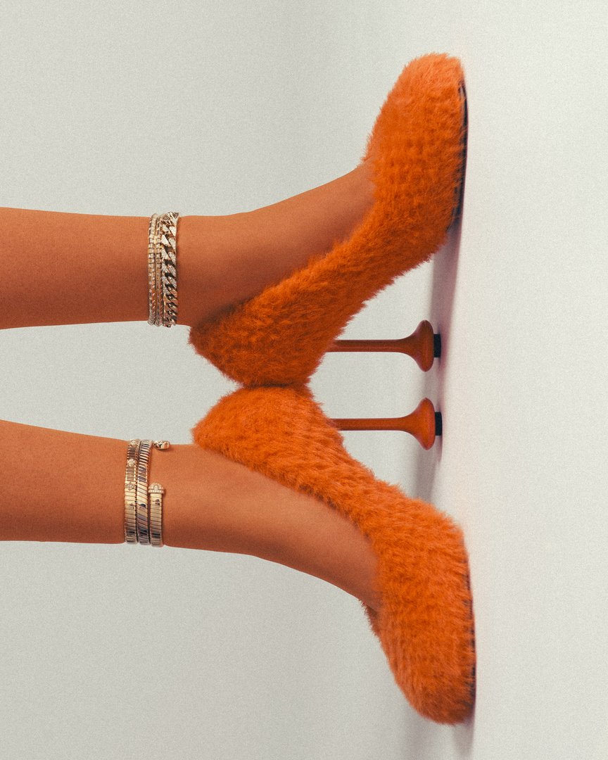Footwear fashion shot: Woman's lower legs with orange fuzzy high-heeled shoes and stacked bracelets against a white wall background.