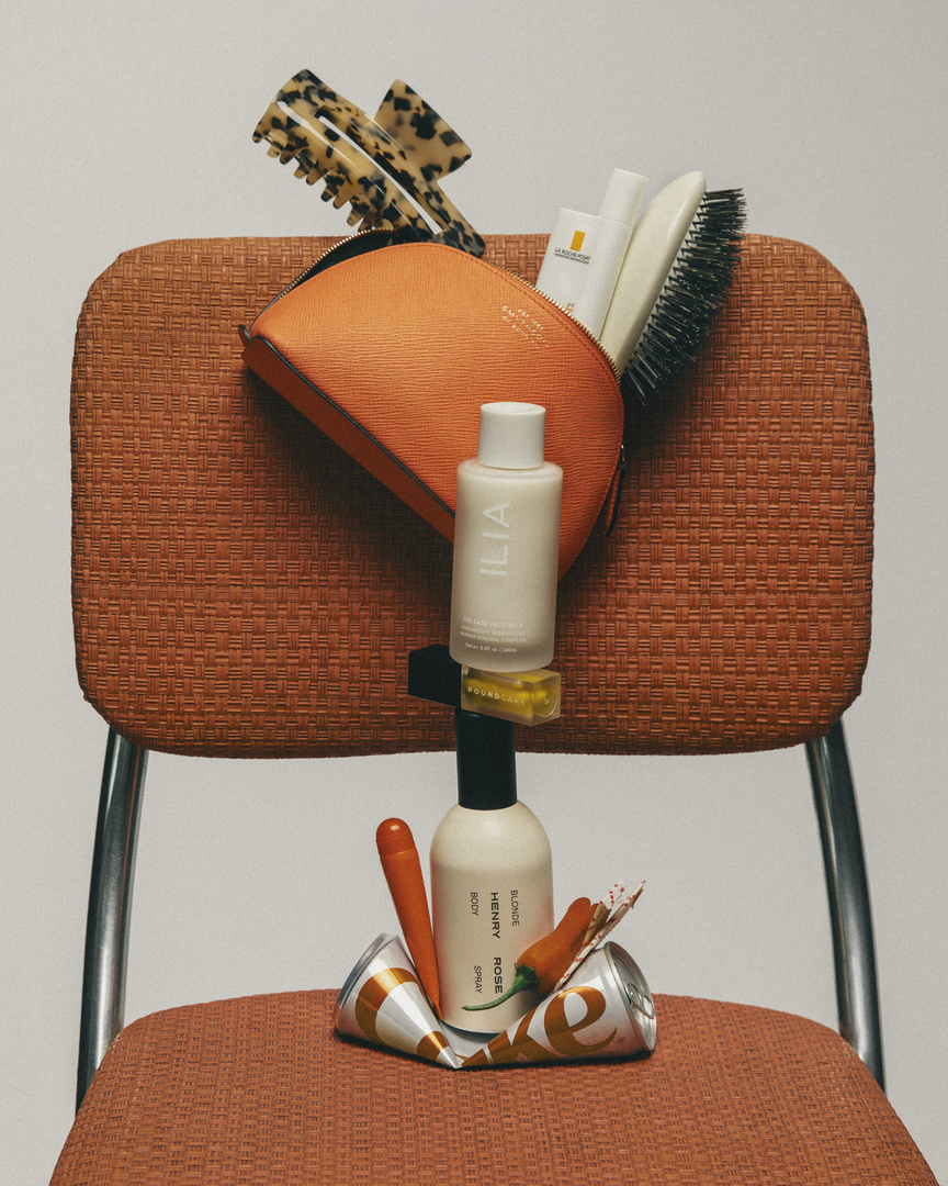 A variety of beauty and cosmetic product bottles and accessories are arranged on an orange chair in a still-life product shot.