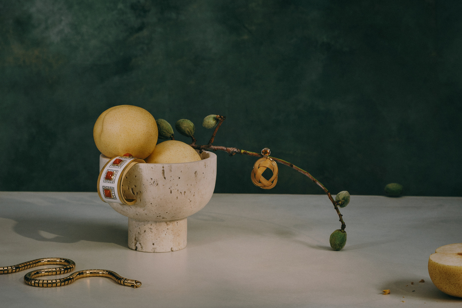 Still life of Asian pears and a green almond branch in a concrete bowl with a gold snake bracelet on a white table against a dark green background.