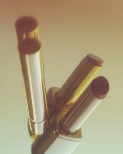 Three lipsticks in gold and white tubes stand against a soft gradient background, showcasing beauty and cosmetic products.