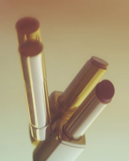 Three lipsticks in gold and white tubes stand against a soft gradient background, showcasing beauty and cosmetic products.
