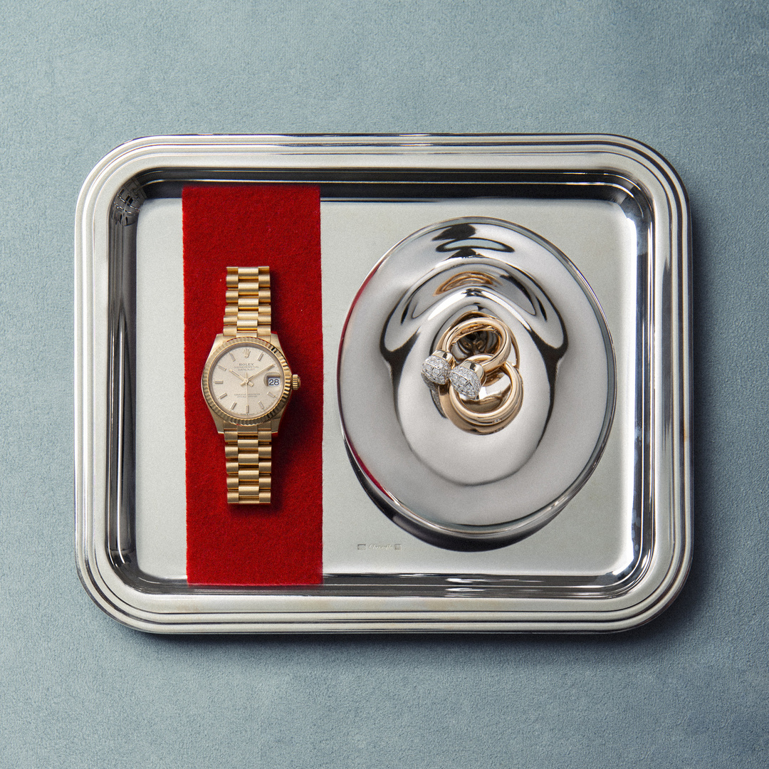 Overhead shot of a gold watch and jewelry on a silver tray with a red velvet accent, set against a solid blue background.