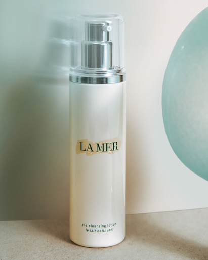 La Mer cleansing lotion in a white bottle with a clear pump top. The bottle sits on a neutral surface with a soft green background.