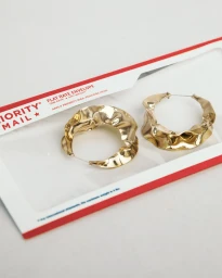 Two textured gold hoop earrings resting inside an open USPS Priority Mail flat rate paper envelope on a white surface.