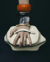 Close-up of a hand resting on a cream handbag, stacked with chunky orange, black, and metallic bangles.