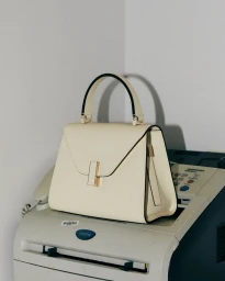 Cream leather handbag with top handle resting on top of a vintage white fax machine in an office corner
