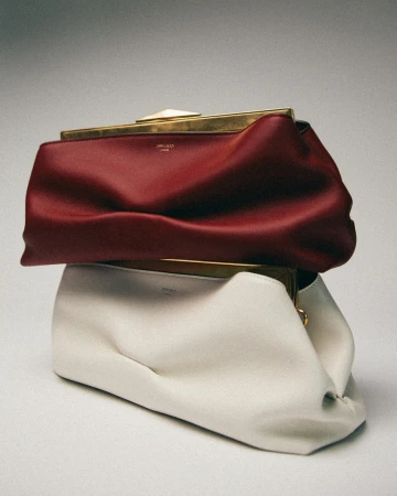 Two stacked leather clutch handbags in burgundy and cream with gold frames against a neutral grey studio backdrop.