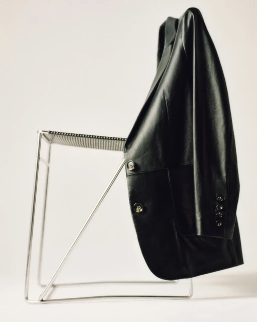 Black leather jacket draped over a modern metal chair against a white backdrop. Clothing item, studio shot, design element.