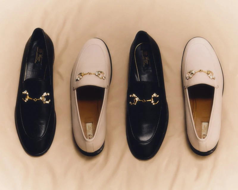 Four stylish leather loafers in black and beige are arranged on a light tan textile background, creating a fashionable footwear display.