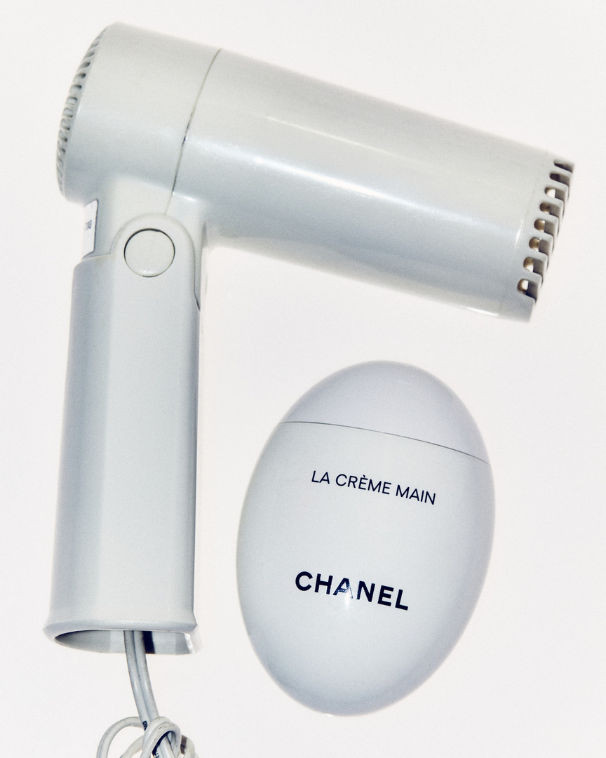 A hair dryer and a white, egg-shaped Chanel hand cream jar sit against a white background in a beauty and personal care product shot.