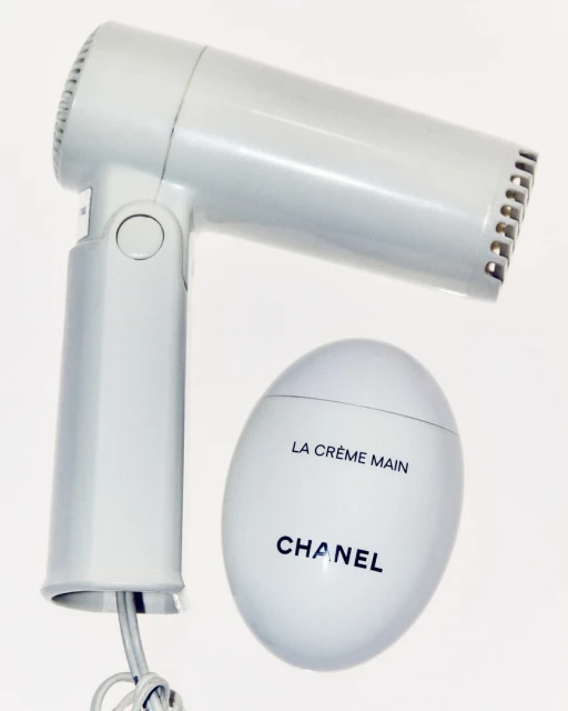A hair dryer and a white, egg-shaped Chanel hand cream jar sit against a white background in a beauty and personal care product shot.