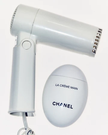 A hair dryer and a white, egg-shaped Chanel hand cream jar sit against a white background in a beauty and personal care product shot.