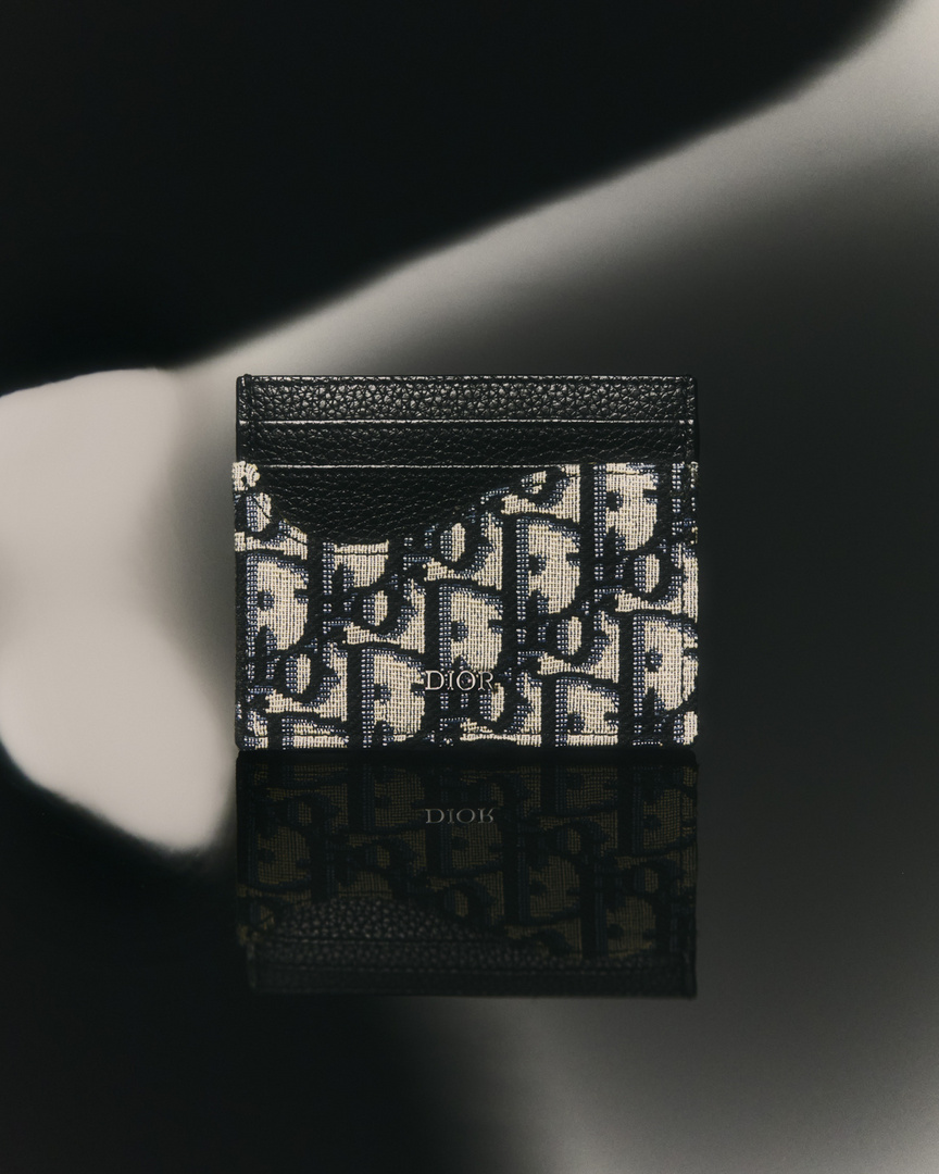 High-angle studio shot of a Dior card holder with an oblique print, reflecting on a glossy black surface against a blurred backdrop.