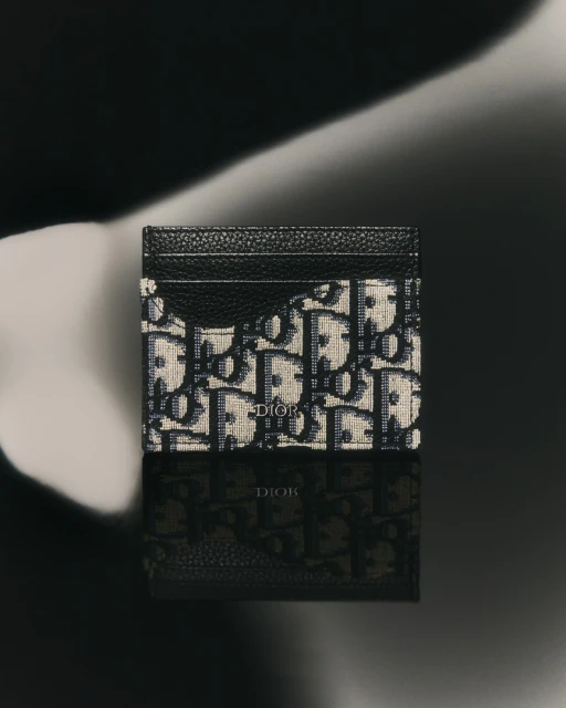 High-angle studio shot of a Dior card holder with an oblique print, reflecting on a glossy black surface against a blurred backdrop.