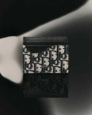 High-angle studio shot of a Dior card holder with an oblique print, reflecting on a glossy black surface against a blurred backdrop.