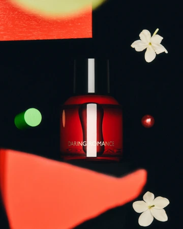 Red perfume bottle with black cap and red/green geometric shapes and white flowers against a dark background in a studio product shot.