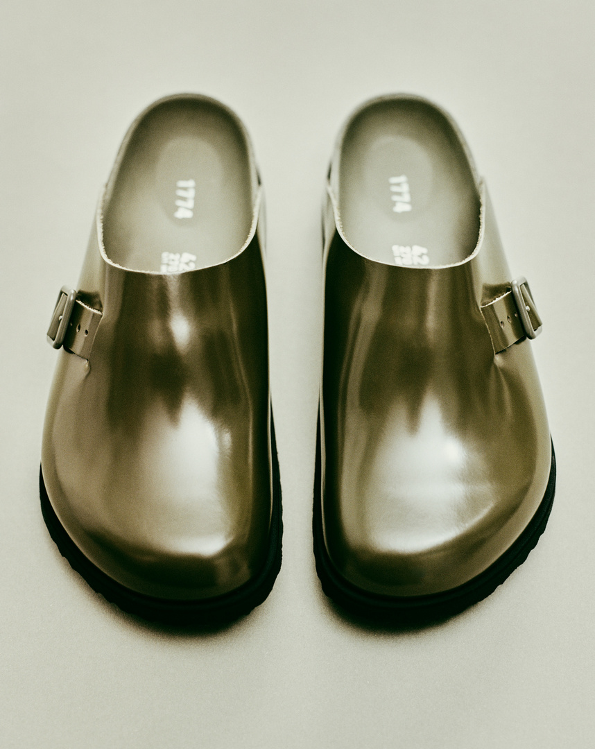 Close-up product shot: a pair of glossy silver leather clogs with black soles, displayed on a seamless, light-colored background.