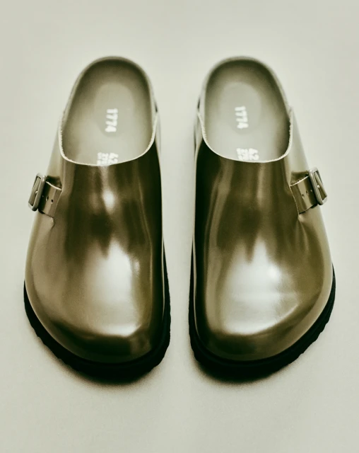 Close-up product shot: a pair of glossy silver leather clogs with black soles, displayed on a seamless, light-colored background.