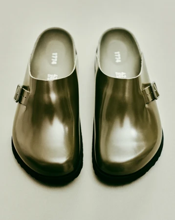 Close-up product shot: a pair of glossy silver leather clogs with black soles, displayed on a seamless, light-colored background.