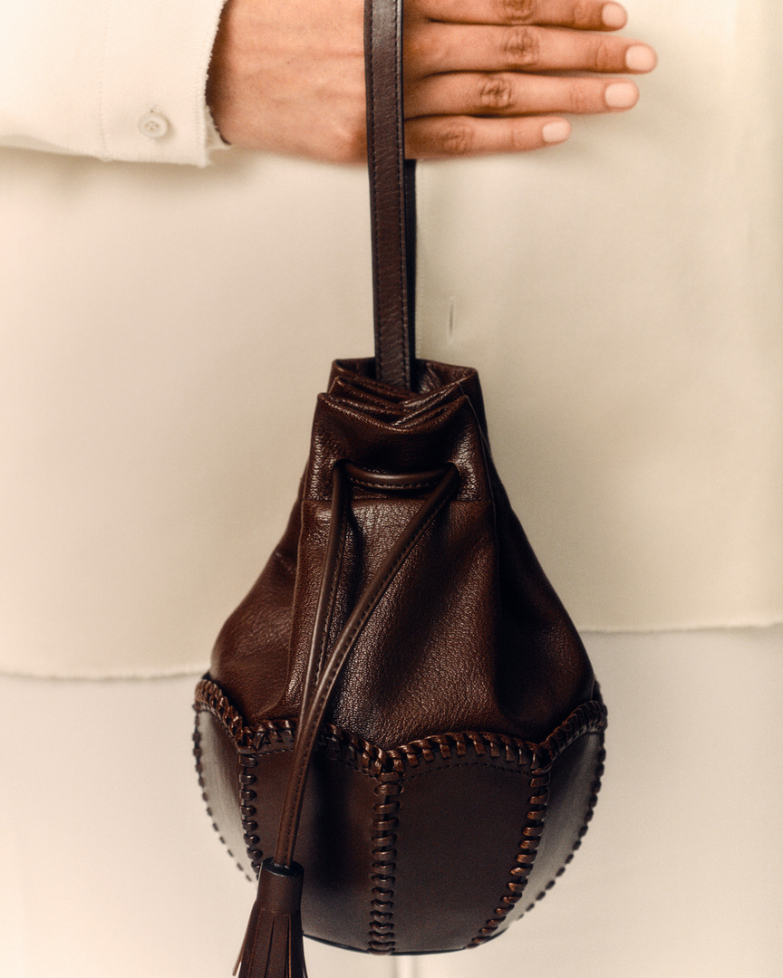 Close-up of a brown leather bucket bag with a drawstring closure held in a person's hand against a white background.