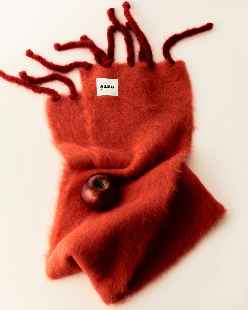 Close-up of a textured reddish-brown fringed scarf with an apple resting on it, set against an off-white backdrop.