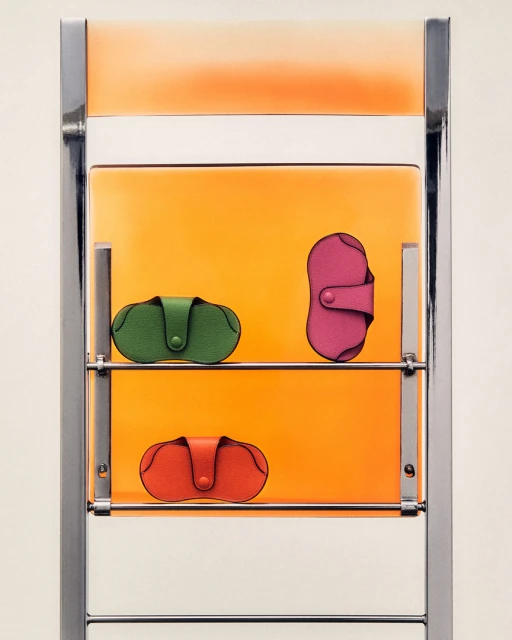 Three colorful leather pouches are presented on a chrome metal shelf against an orange backdrop in a minimalist composition.