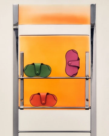 Three colorful leather pouches are presented on a chrome metal shelf against an orange backdrop in a minimalist composition.