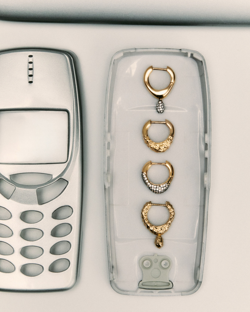 Gold hoop earrings displayed in a transparent plastic case alongside a vintage silver-colored cell phone on a plain white background.