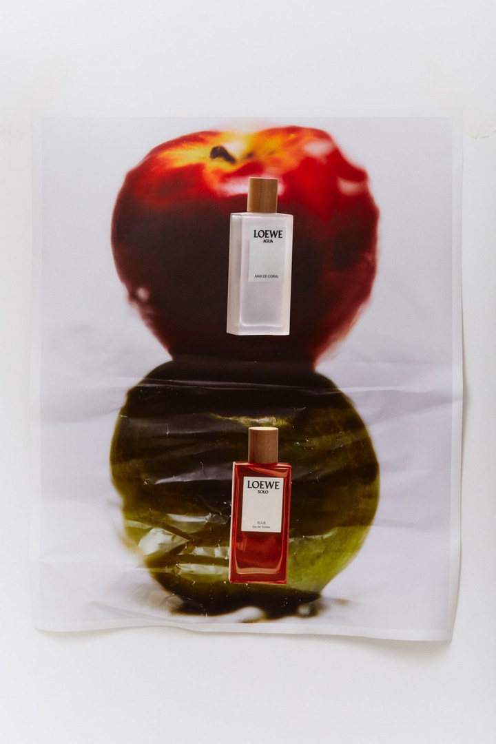 Two Loewe Solo fragrances are arranged over a printed apple image, one bottle reflecting in a glossy surface, studio lighting.