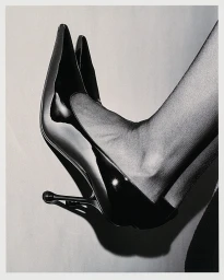 Monochrome close-up of patent leather stiletto pumps worn with sheer stockings by Eduard Sanchez Ribot.