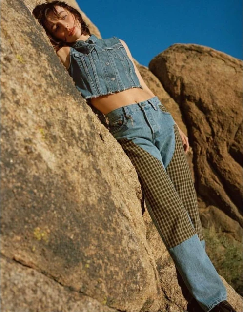 A woman in a denim outfit leans against a rock formation under a clear blue sky in an outdoor setting. Fashion shot.