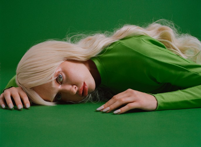 Young woman with long blonde hair and light skin, wearing a green top, laying on a green background, looking at the camera.