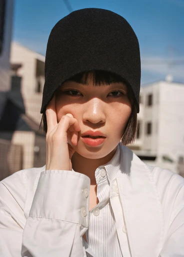 Young Asian woman with short hair wearing a black cloche hat and white button-down shirt poses outdoors in an urban setting on a sunny day.