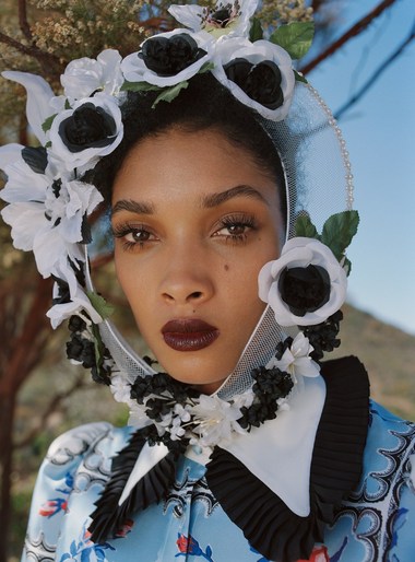 Close-up portrait of an adult female wearing a floral headpiece with a ruffled collar, set against an outdoor mountain background.