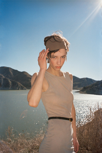 Woman in a beige dress and matching headband adjusting her hair outdoors near a lake and mountains under a clear sky.