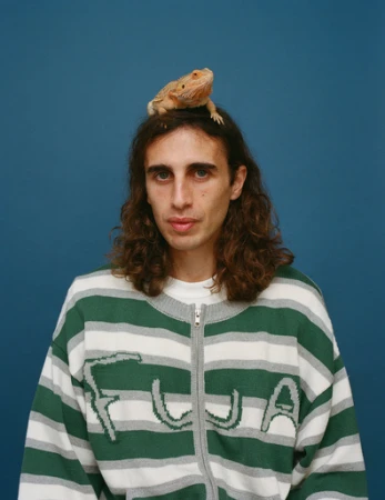 A young adult with long brown hair poses indoors with a bearded dragon on his head, wearing a striped sweater against a solid blue backdrop.