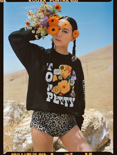 Young woman in a black sweatshirt and shorts poses outdoors surrounded by colorful flowers, styled with face-framing braids and flower details.