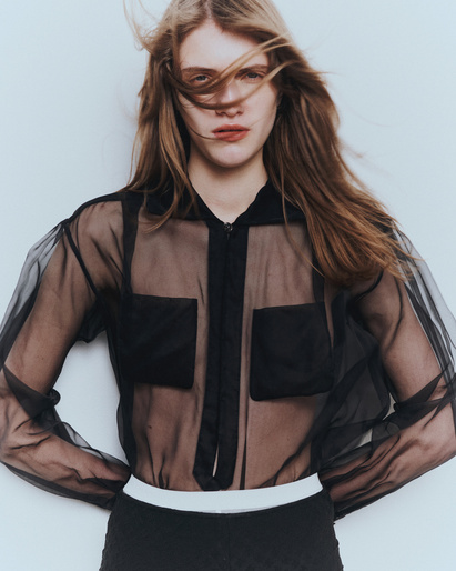 Model with long brown hair blowing in the wind wearing a sheer black button-up blouse against a white background