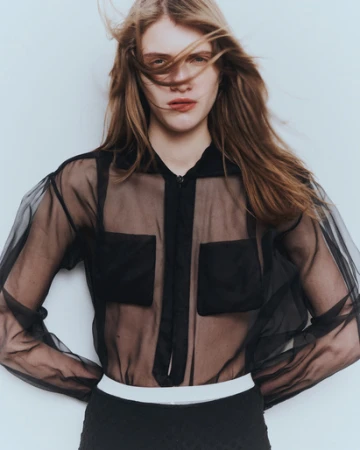 Model with long brown hair blowing in the wind wearing a sheer black button-up blouse against a white background