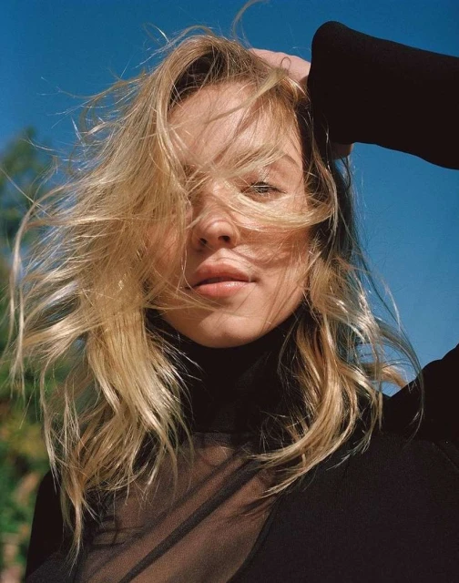 Close-up portrait of a young woman with windswept blonde hair against a bright blue sky, wearing a sheer black turtleneck top.