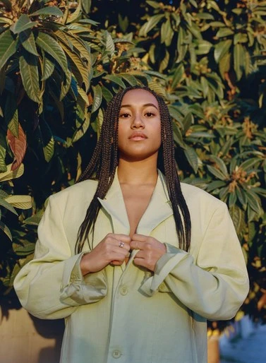 Woman in a light-green oversized jacket poses outdoors with a neutral expression against a leafy green background in soft, diffused light.