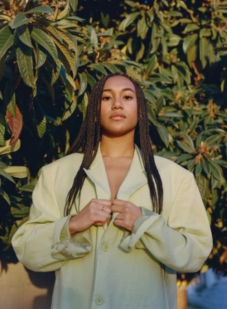 Woman in a light-green oversized jacket poses outdoors with a neutral expression against a leafy green background in soft, diffused light.