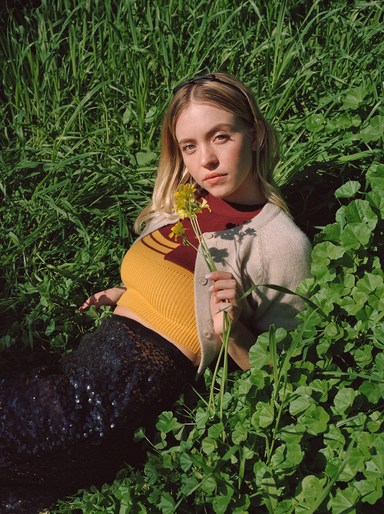 Blonde woman in yellow top and sequin skirt lies in a green field, holding yellow flowers, looking at the camera.