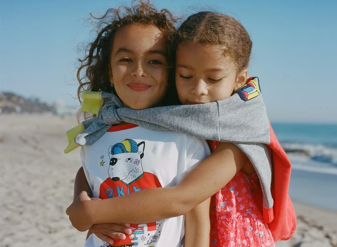 Two young girls hugging on a sandy beach on a sunny day. One child wears a dog graphic tee shirt with a grey hooded sweatshirt.