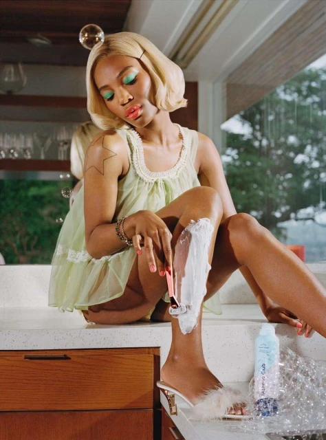 A woman shaves her leg with shaving cream and a razor while sitting on a marble counter in a bright, indoor environment.