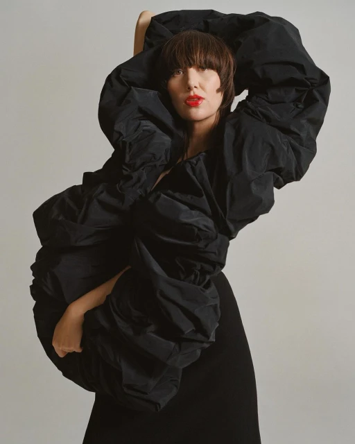 Woman in extravagant black jacket poses against a grey backdrop, showcasing fashion and studio photography with bright red lipstick.