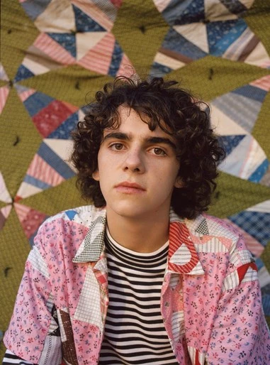 Portrait of a young adult with curly brown hair wearing a patchwork shirt and black and white striped top, in front of a patterned quilt.