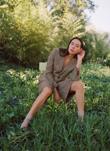 Aubrey Plaza sitting on a chair in a field, wearing a tan suit with short shorts, surrounded by green foliage in an outdoor setting.
